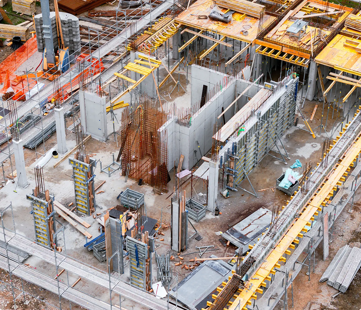 Aerial view of a building construction site with exposed concrete walls, steel reinforcements, scaffolding, and various construction materials scattered around.