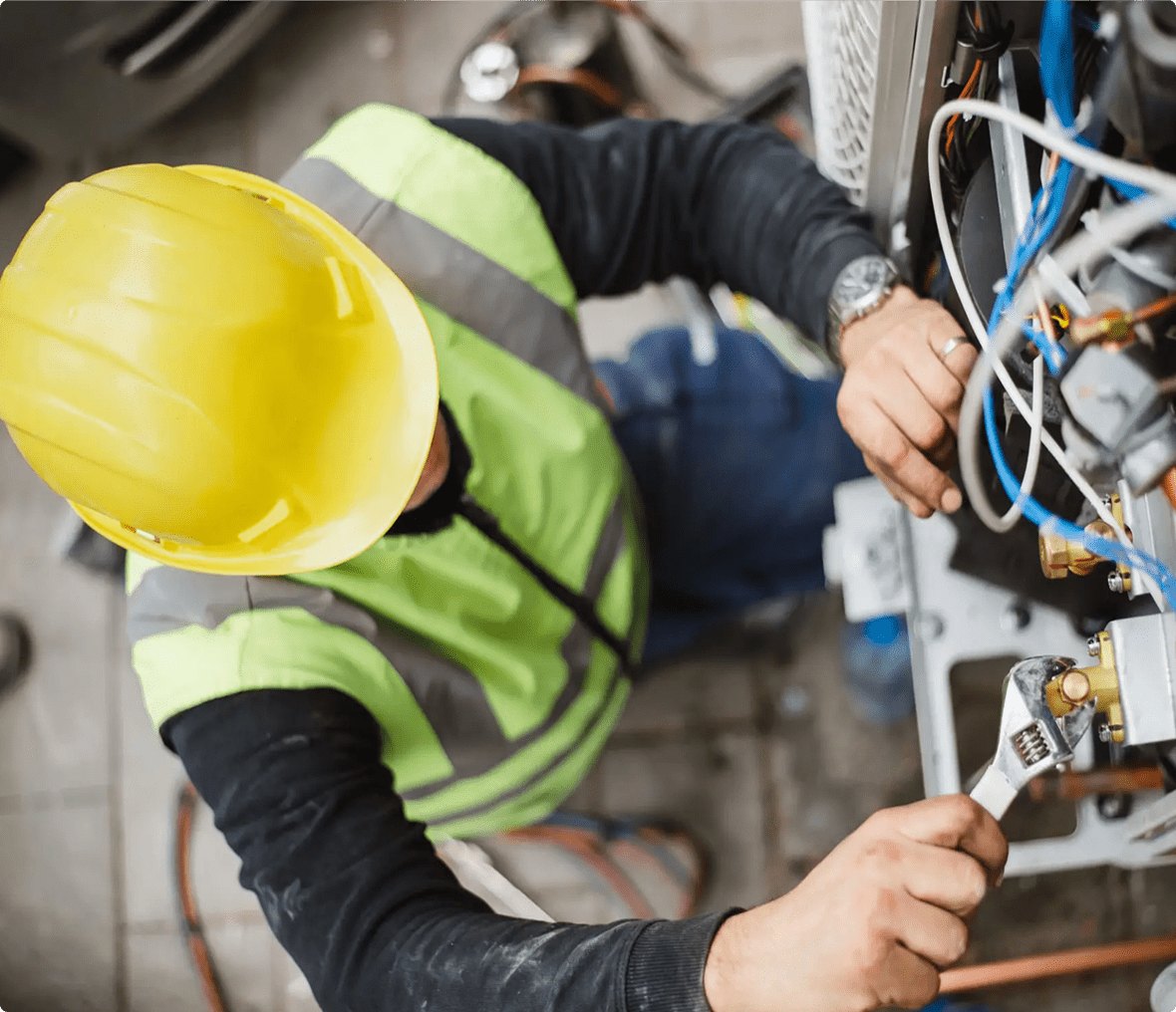 Person wearing a yellow hard hat and reflective vest uses a wrench to work on electrical or mechanical equipment with connected wires.