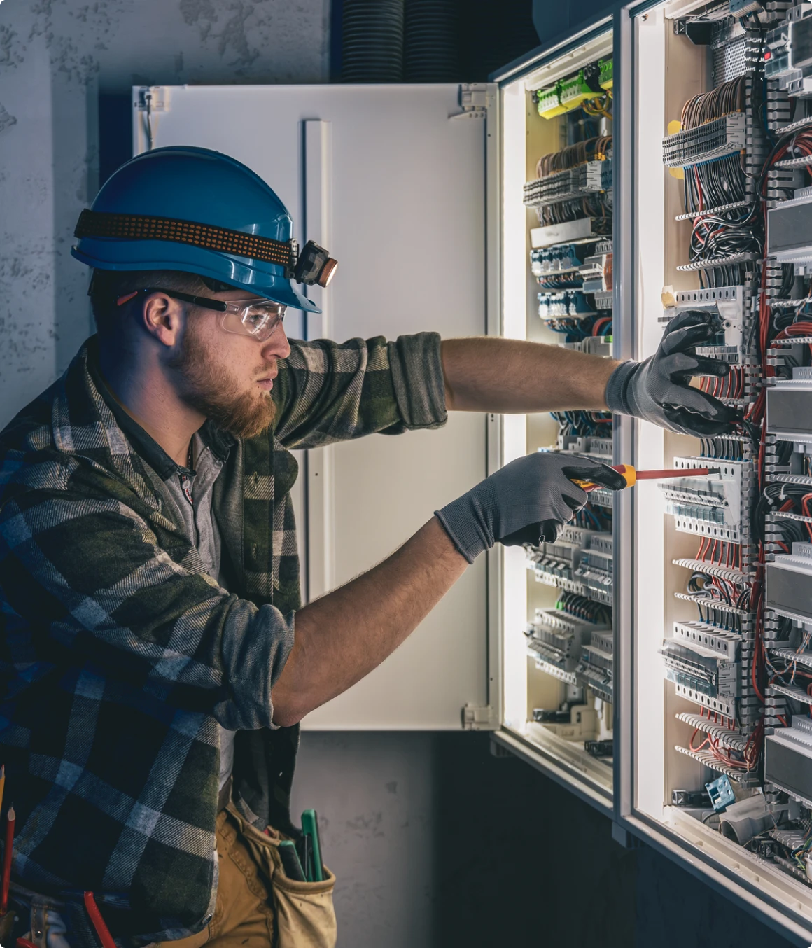An electrician wearing a helmet, gloves, and safety glasses uses a screwdriver to work on wires inside an open electrical panel.