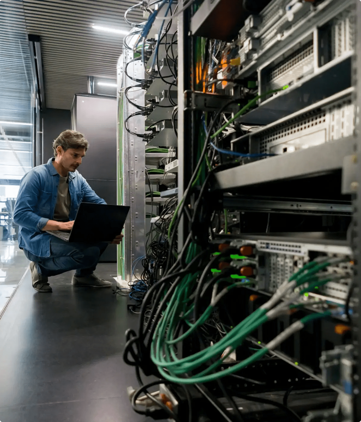 A person kneels in a server room, using a laptop next to server racks filled with network cables and computer equipment.