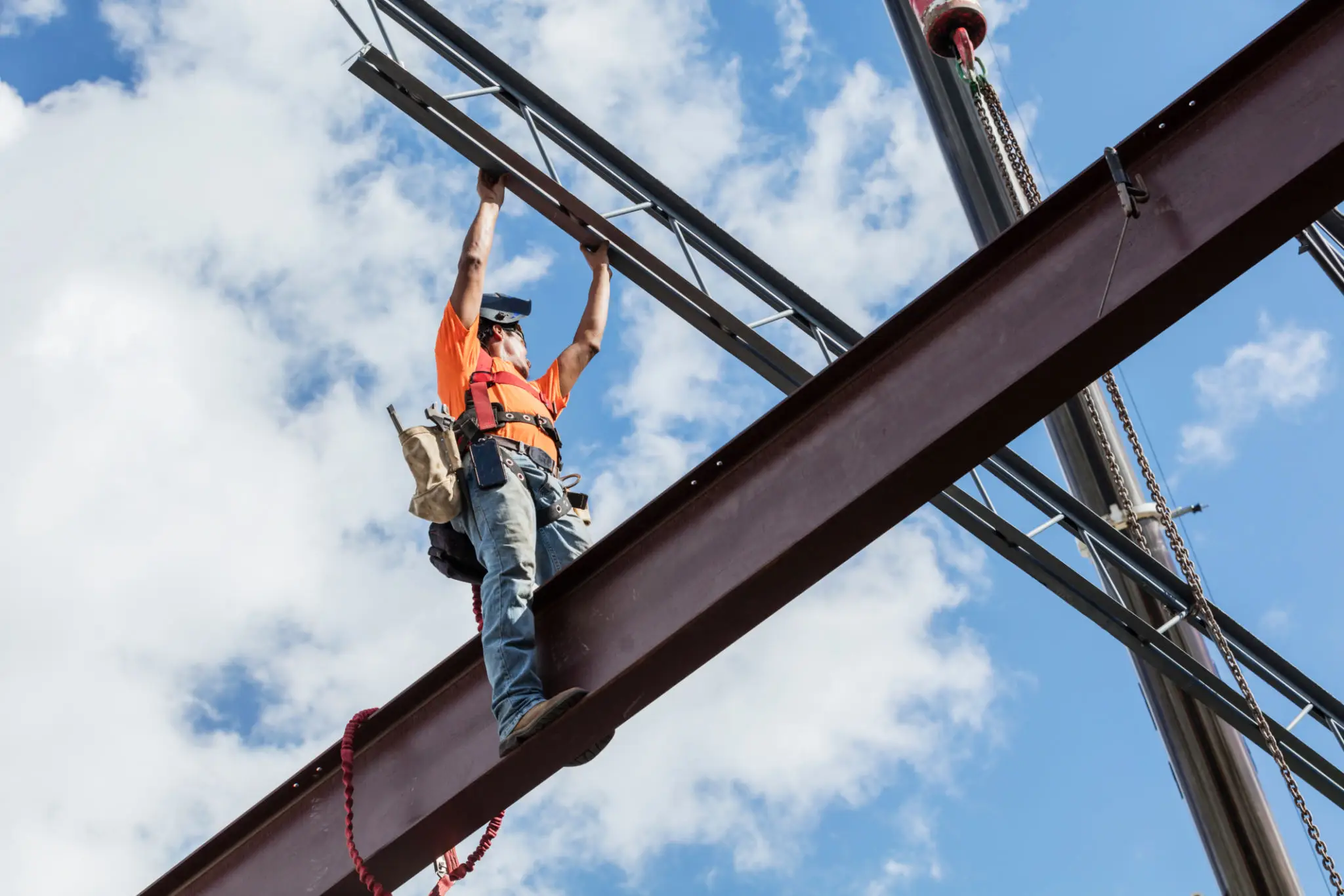 A construction worker in a safety harness stands on a steel beam, guiding a metal structure into place against a partly cloudy sky.