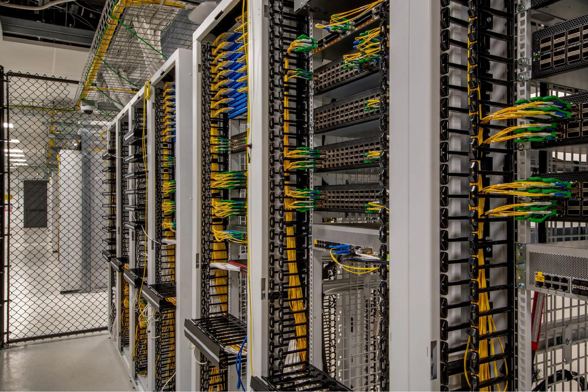 Rows of server racks in a data center with organized yellow, blue, and green network cables connected to patch panels, enclosed by a chain-link fence.