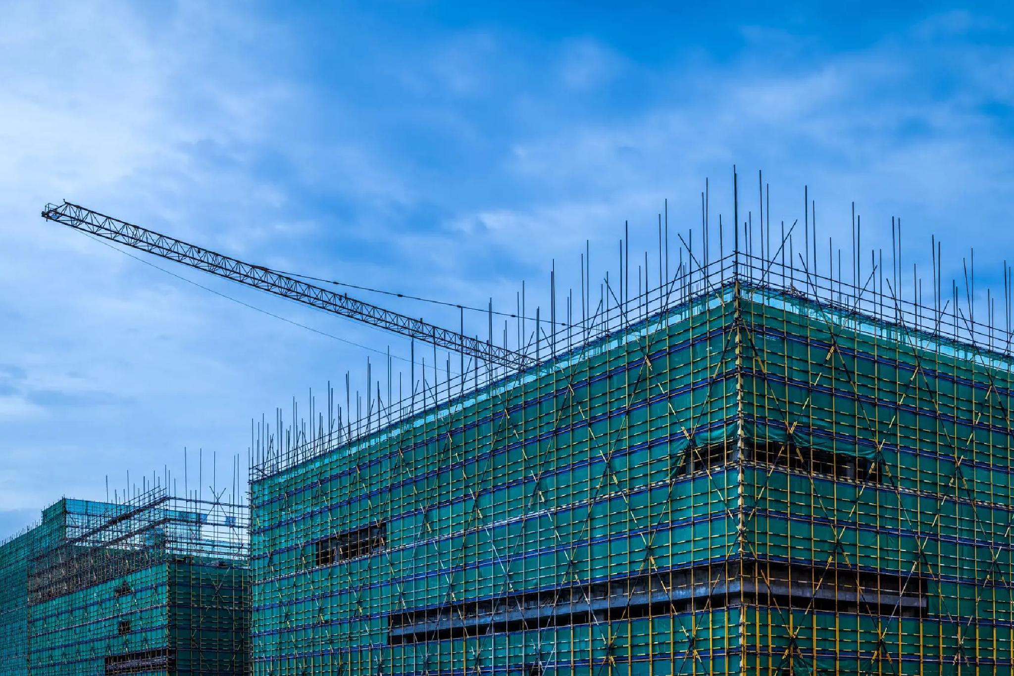 Two buildings under construction with green scaffolding and exposed rebar, with a crane arm extending over one building, set against a partly cloudy blue sky.