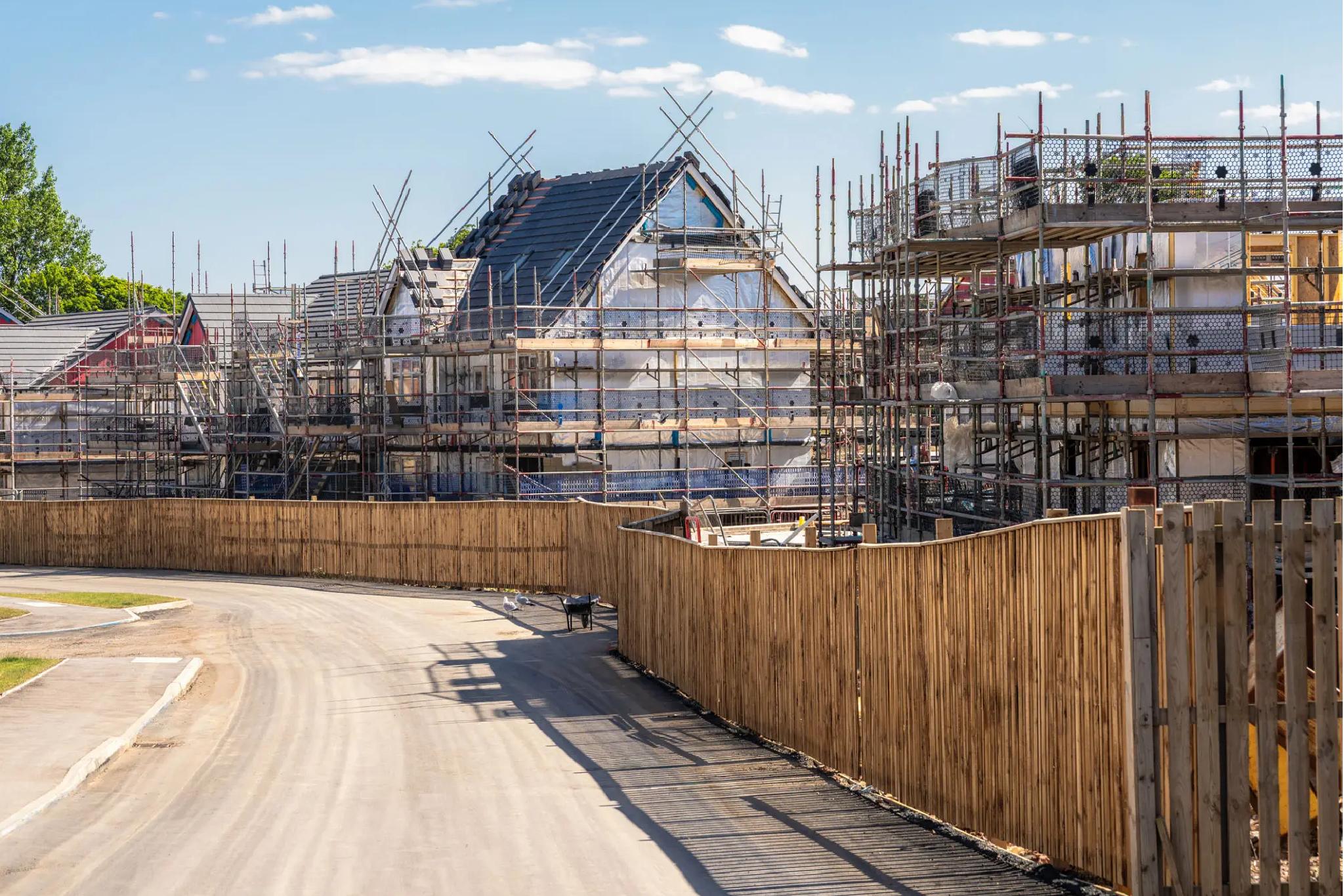 Several houses under construction, covered with scaffolding and partially built, behind a wooden fence along a curved road on a sunny day.