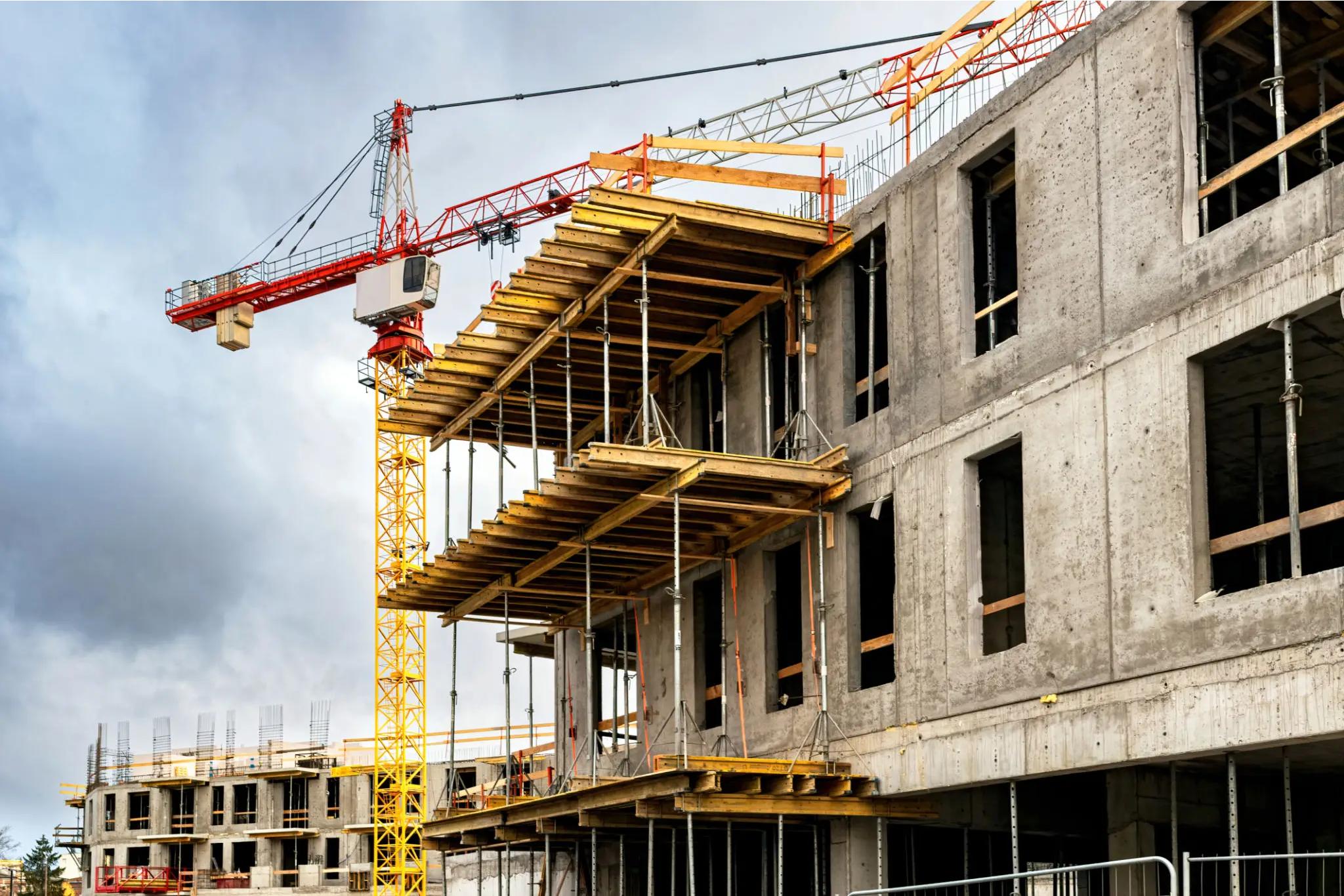 Concrete building under construction with exposed floors, scaffolding, and a yellow tower crane against a cloudy sky.