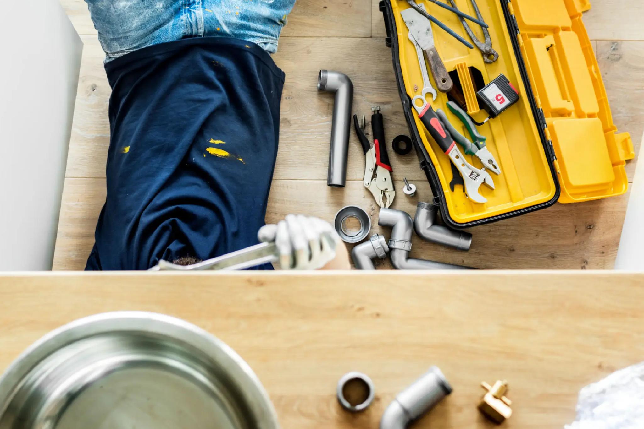Person in paint-stained clothes working under a sink with a wrench; plumbing tools, pipes, and an open yellow toolbox are on the floor.