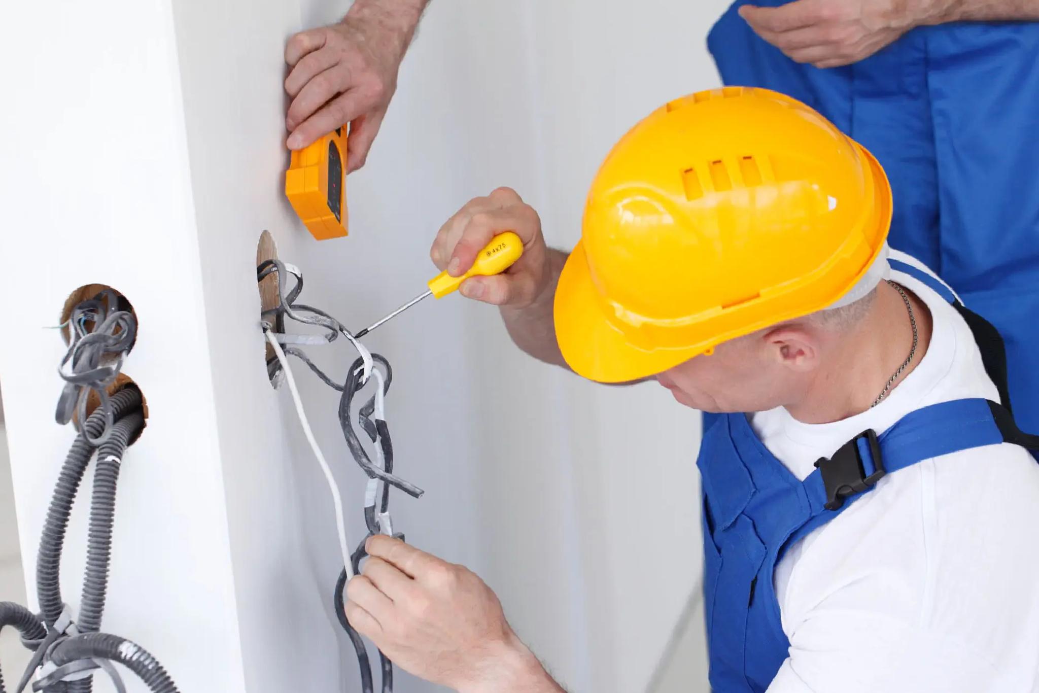 An electrician in a yellow hard hat uses a screwdriver to work on exposed electrical wires in a wall while another person holds a testing device.