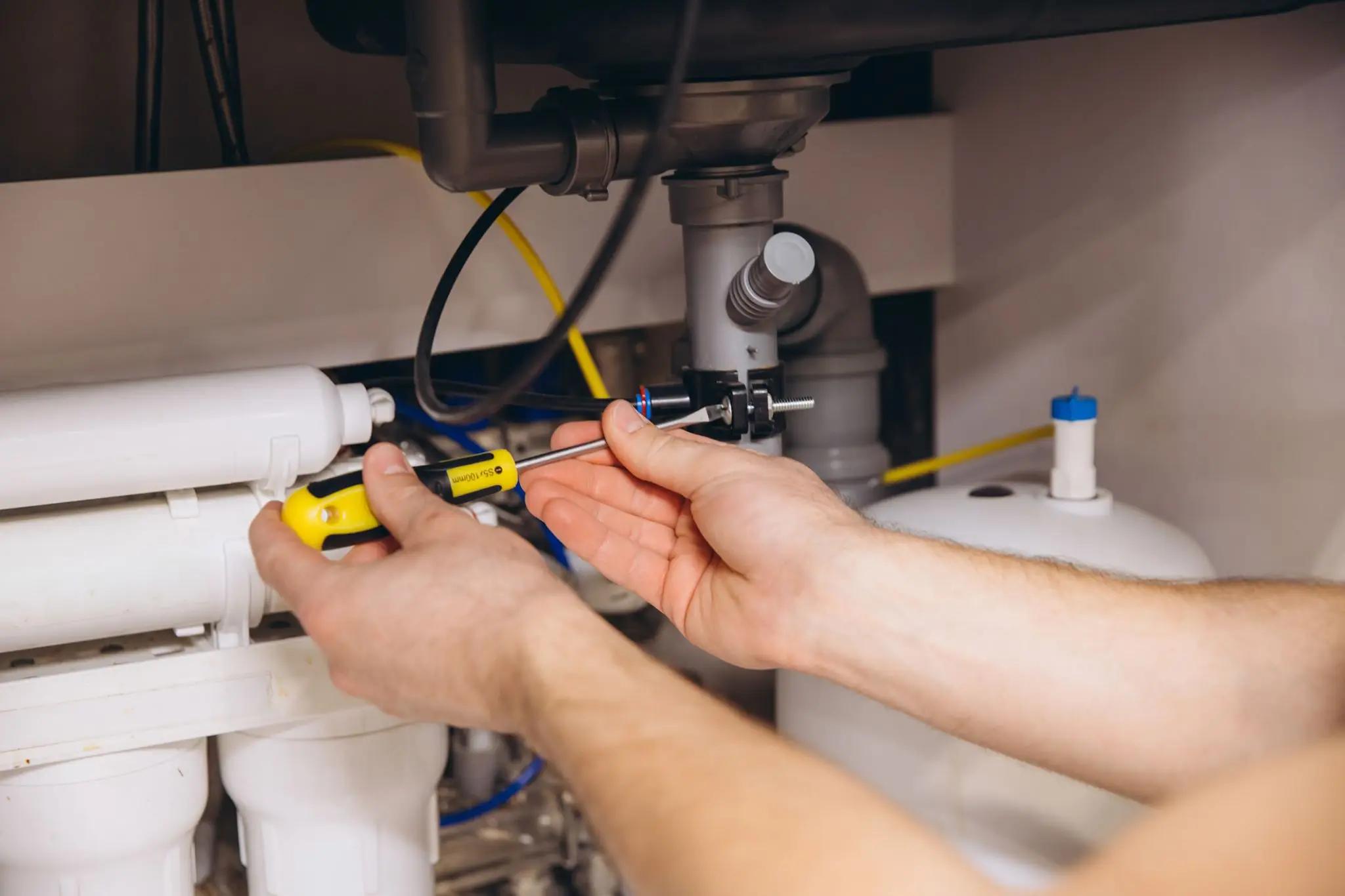 Person using a screwdriver to adjust or repair plumbing connections under a sink, near water filtration equipment.