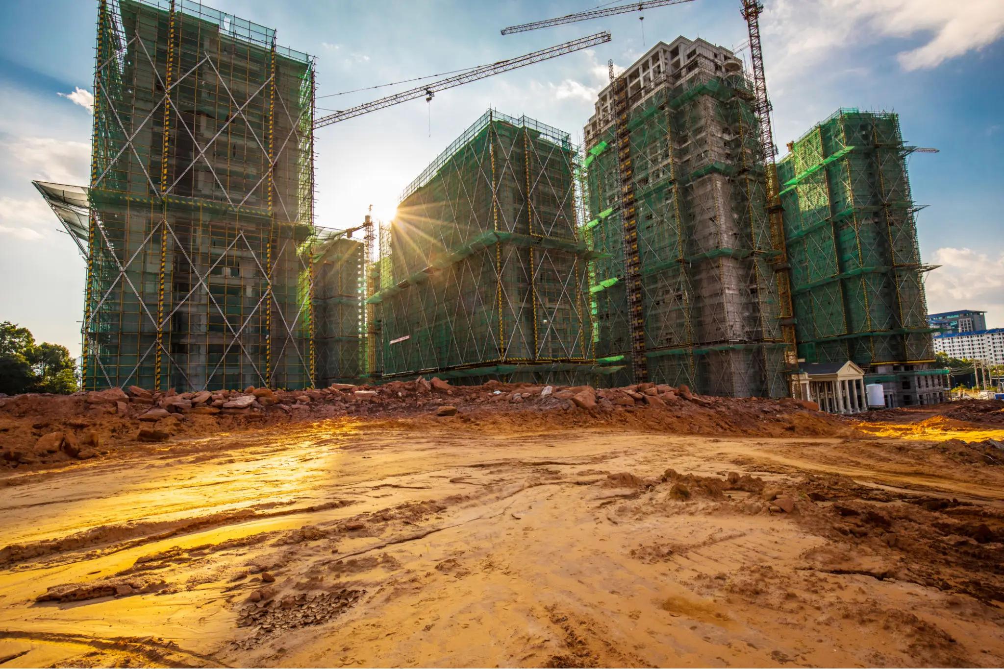 High-rise buildings under construction with scaffolding and green netting, surrounded by dirt and construction materials, as the sun sets in the background.