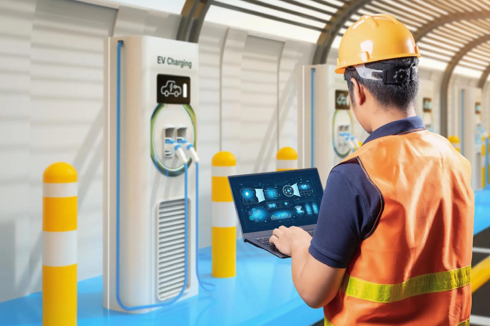 A worker in a hard hat and safety vest uses a laptop to monitor electric vehicle charging stations in a modern indoor facility.