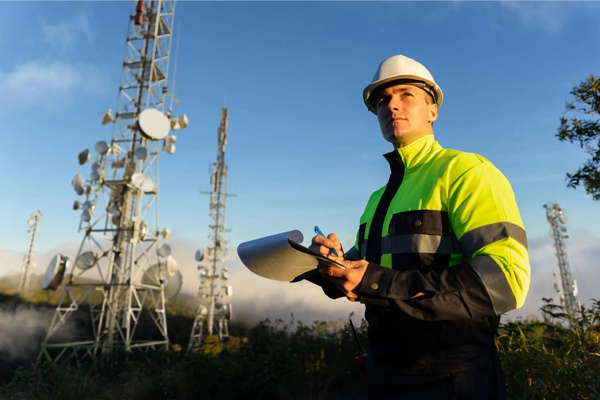 A worker in a high-visibility jacket and hard hat writes on a clipboard near several telecommunications towers outdoors.
