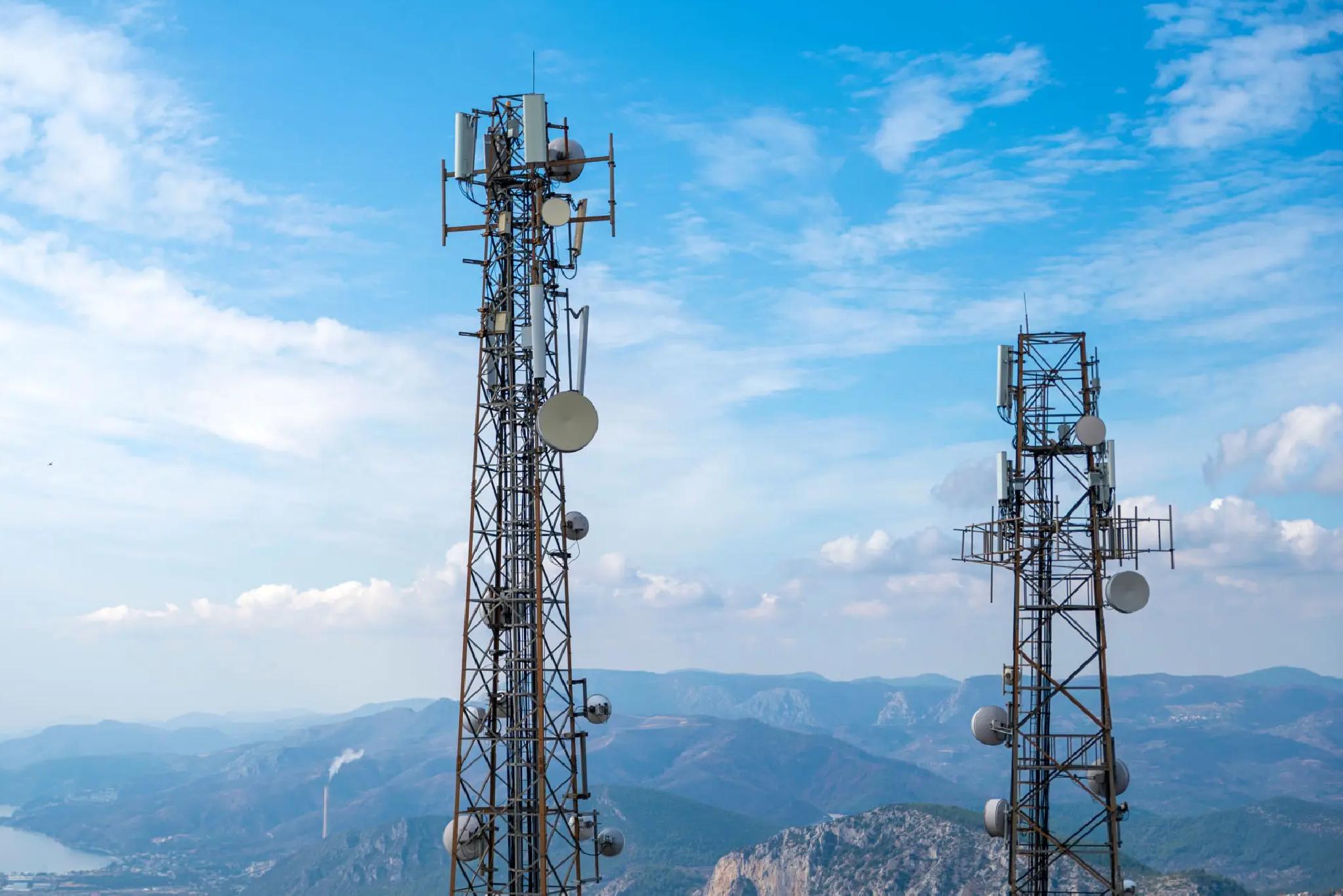 Two tall telecommunications towers with antennas and satellite dishes stand on a hilltop, overlooking mountains under a partly cloudy blue sky.