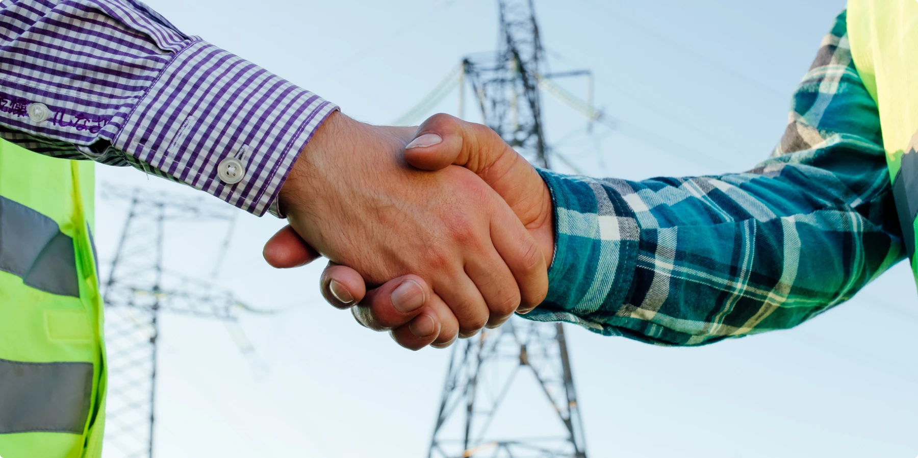 Two people wearing checked shirts and safety vests shake hands outdoors, with an electricity transmission tower in the background.