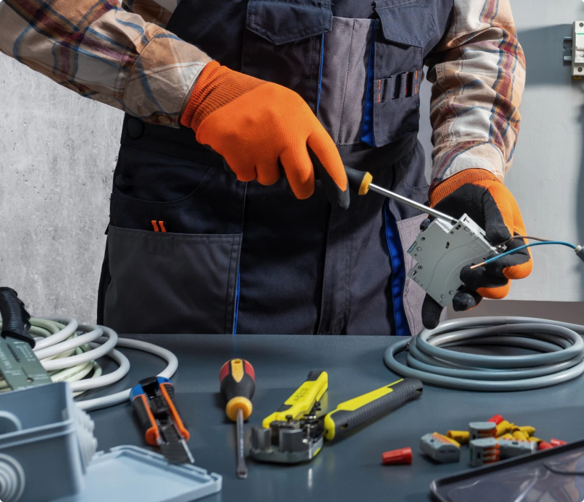 Electrician wearing orange gloves uses a screwdriver to work on an electrical outlet, with various tools and cables on a table in front.