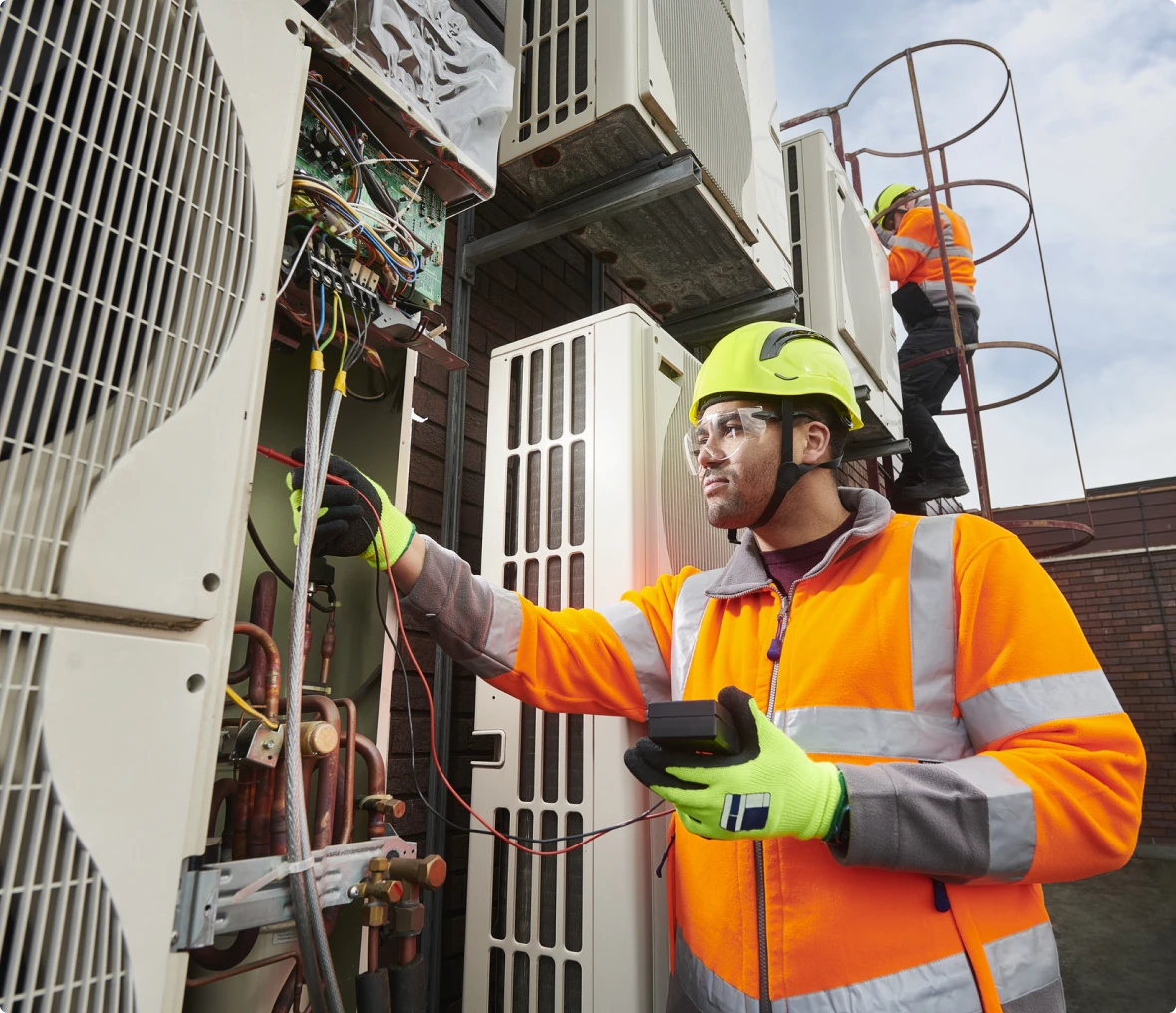 Two workers in high-visibility clothing and helmets service large HVAC units outdoors; one checks wiring with a meter, while the other climbs a metal ladder in the background.