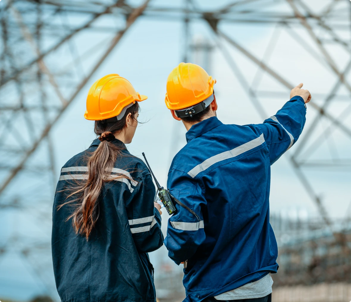 Two workers wearing orange helmets and blue uniforms stand outdoors; one holds a walkie-talkie while the other points toward a metal power structure in the background.