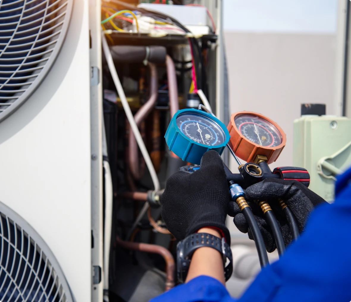 Technician wearing gloves uses pressure gauges to check an HVAC system outdoors, with pipes and electrical components visible in the background.