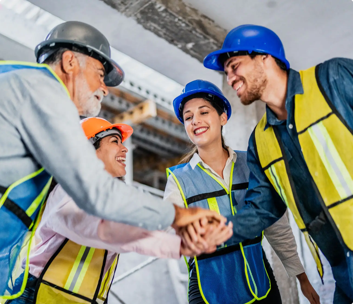 Four construction workers in safety vests and helmets stand together indoors, smiling and stacking their hands in a gesture of teamwork.