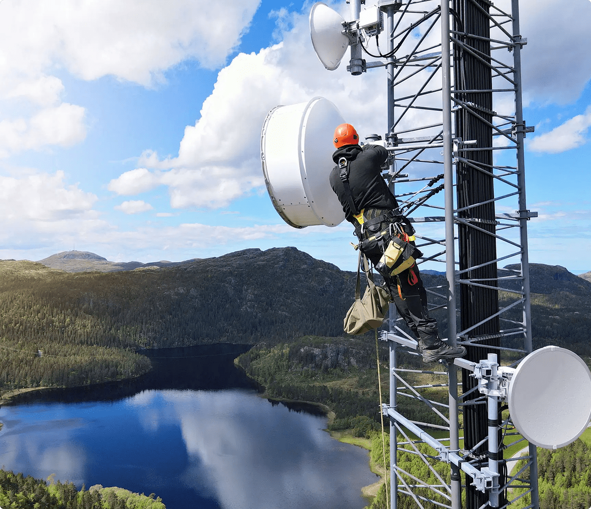 A worker in safety gear climbs a communications tower above a lake and forested landscape, performing maintenance on large white antennas.